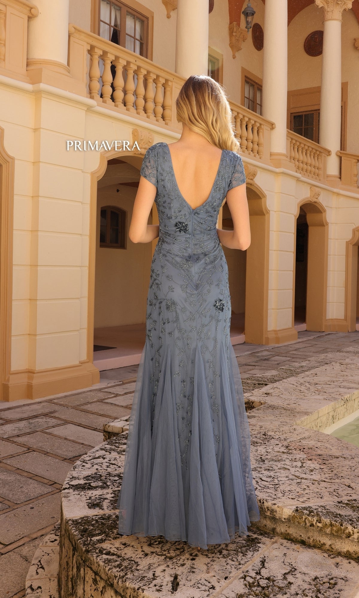 A woman wearing the Long Prom Dress 13112 by Primavera, a light blue floral beaded mermaid gown with short sleeves and an open back, stands outdoors before a beige building with arches and columns.